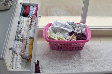 clothes basket on the floor and open drawer full of folded baby's clothes. Basket on white carpet next to a window. Dry clothes after laundry time