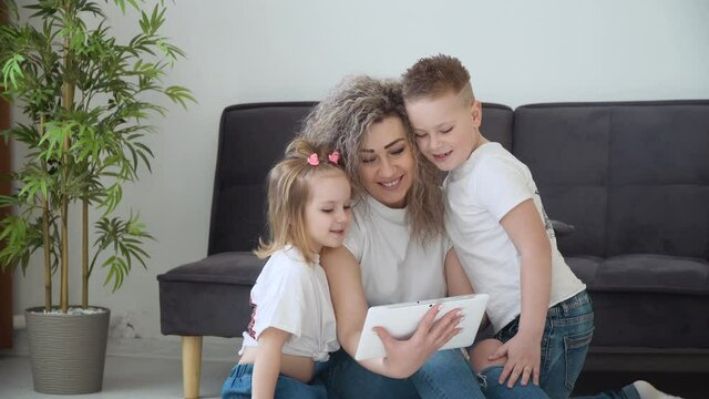 Mom and daughter and son use the tablet together in a cozy living room. The family is dressed in the same basic white T-shirts. Leisure with children. Family entertainment