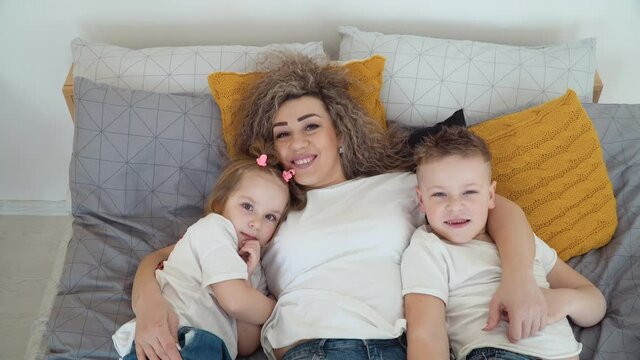 Mom and daughter and son lie in bed together hugging. Mom and children are sleeping and suddenly open their eyes and look at the camera. The family is dressed in the same basic white T-shirts