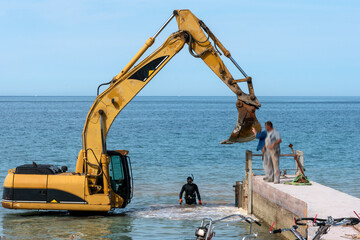 Diver controls the work of a excavator spreading gravel in shallow water in the sea at the promenade