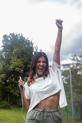 vertical a photo of happy latina woman outside with headphones and cell phone celebrating with one hand up. Listening to music, video calling, working remotely.