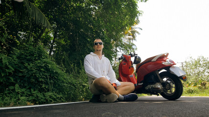 Man on red motorbike in white clothes drive on forest road trail trip. One men caucasian tourist in sunglasses sit on pavement, relax, near scooter.