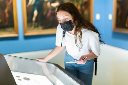 Focused young girl in a protective mask, visiting a museum during a pandemic, looks at the exposition behind a glass..case, holding an information booklet