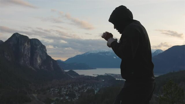 Adult Middle Age Caucasian Man wearing a black hoodie practicing shadow boxing on top of a mountain. Sunset Sky. Taken in Squamish, North of Vancouver, British Columbia, Canada. Slow Motion