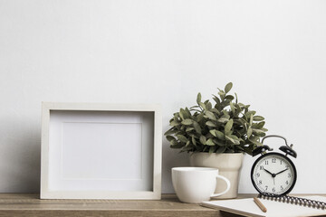 A white photo frame with plant, coffee cup and clock on table