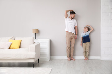 Father and son comparing their heights near wall in living room - obrazy, fototapety, plakaty