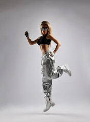 Fototapeta na wymiar Athletic woman, sporty girl in brutal shoes, silver hip-hop lose fit pants, black top and sunglasses is jumping, exercising in gym on gray background