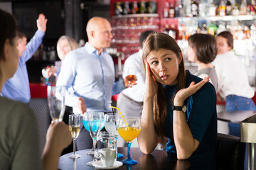 Upset young woman sitting alone at corporate bar party on background with cheerful colleagues..