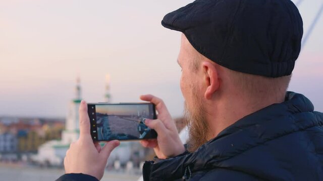 A bearded man in a cap. Side view in profile of the face. He takes a photo with a smartphone from the bridge as the ice moves on the river. In the background in blur the evening city.