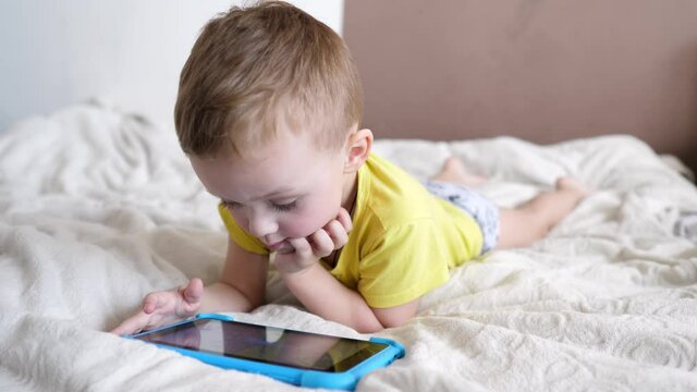 Children's technical addiction. Little preschool boy using digital tablet device lying on carpet floor alone. A small child is holding a computer surfing the Internet while playing a game at home