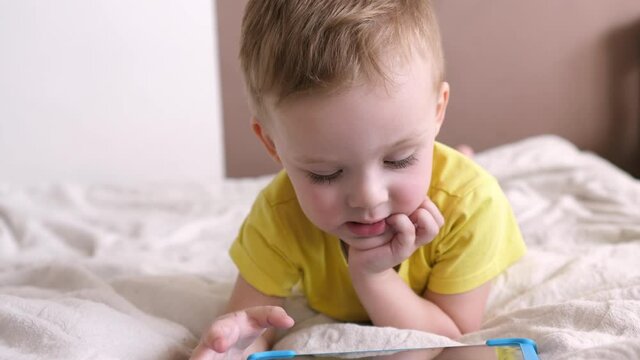 Children's technical addiction. Little preschool boy using digital tablet device lying on carpet floor alone. A small child is holding a computer surfing the Internet while playing a game at home