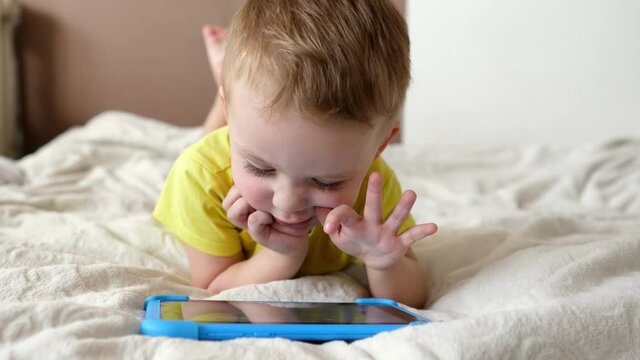 Children's technical addiction. Little preschool boy using digital tablet device lying on carpet floor alone. A small child is holding a computer surfing the Internet while playing a game at home