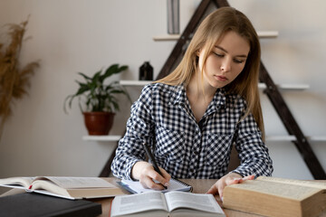 Motivated student sits with textbooks at a desk studying at home. A girl writes homework on mathematics in a notebook or a teacher prepares for a lecture in college remotely online.
