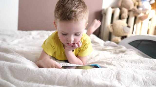 Children's technical addiction. Little preschool boy using digital tablet device lying on carpet floor alone. A small child is holding a computer surfing the Internet while playing a game at home