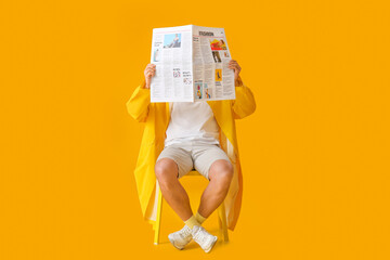 Stylish young man with newspaper sitting on chair against yellow background
