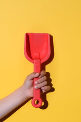 red plastic shovel on the hands of an unrecognizable child hand. It is projecting a deep shadow on yellow background. Summer concept