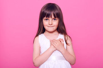 Charming honest little girl wearing white dress looking at camera, keeping hands on chest, has...