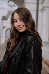 Young woman in black leather jacket and sweater standing in front of white wall looking at camera. Urban style and street fashion. Smiling girl with curly hairstyle in casual outfit