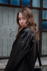 Smiling young woman in black leather jacket standing in front of white wall looking at camera over shoulder. Urban style and street fashion. Girl with curly hairstyle in casual outfit