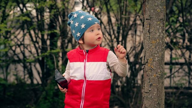 A boy in a blue cap and a red jacket holds a smartphone in his hands and plays with a dry tree.