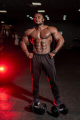 Fototapeta na wymiar Halfnaked strong african american bodybuilder standing with sport equipment in dark modern gym.