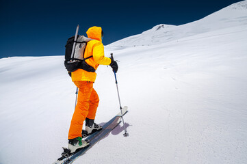Young male freerider guide in a yellow ski suit with a backpack on his shoulders climbing the mountain on skis on a sunny day.