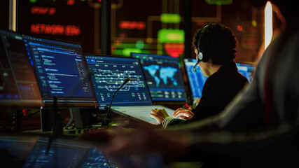 Multiethnic Female IT Technical Support Specialist Talking with a Client in Headphones and Working on Desktop Computer in Monitoring Control Room with Big Digital Screens with Server Blockchain Data.