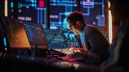 Professional IT Technical Support Specialist Making an Intercom Announcement on a Microphone and Working on Desktop Computer in Monitoring Control Room with Big Digital Screens with Server Data.