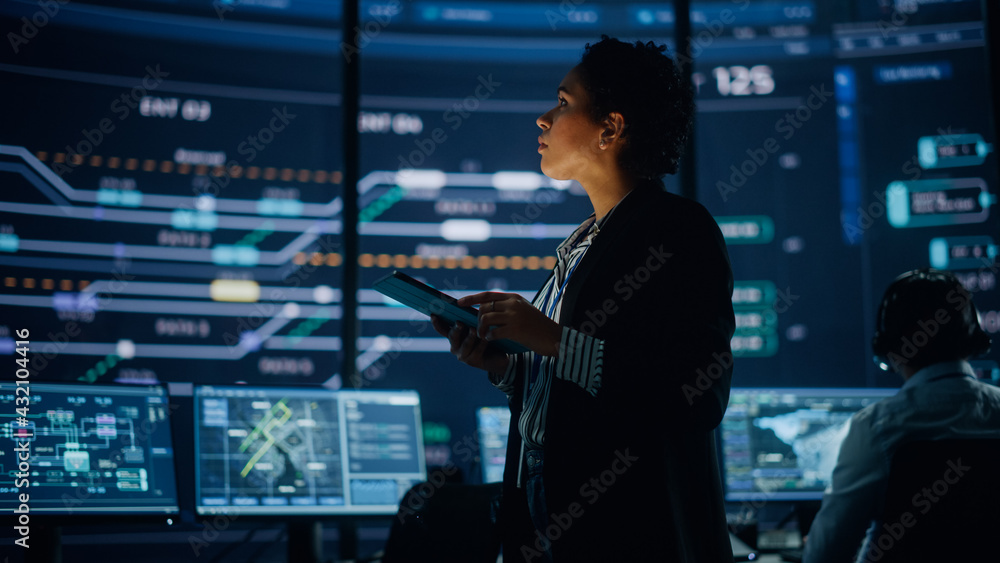 Poster Young Multiethnic Female Government Employee Uses Tablet Computer in System Control Monitoring Center. In the Background Her Coworkers at Their Workspaces with Many Displays Showing Technical Data.