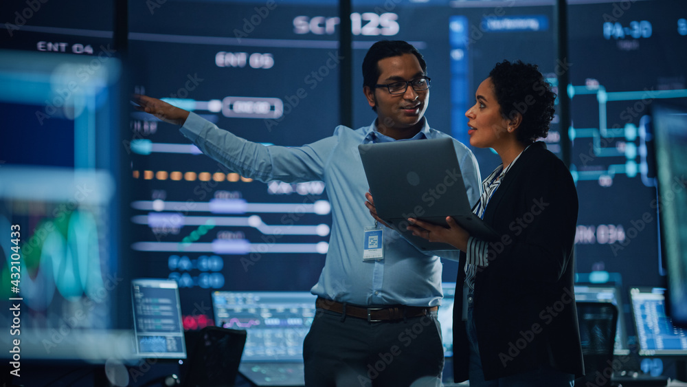 Poster In the System Control Room Project Manager and IT Technical Engineer with Laptop Have Discussion, surrounded by Multiple Monitors with Graphics. Big Monitor Shows Interactive Server Blockchain Info.