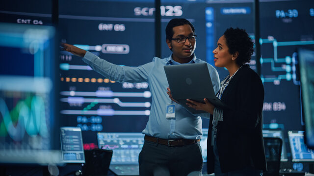 In The System Control Room Project Manager And IT Technical Engineer With Laptop Have Discussion, Surrounded By Multiple Monitors With Graphics. Big Monitor Shows Interactive Server Blockchain Info.