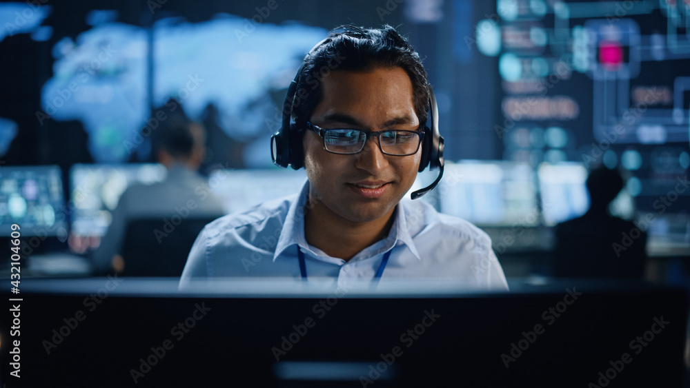 Poster Portrait of Professional IT Technical Support Specialist Working on Computer in Monitoring Control Room with Digital Screens. Employee Wears Headphones with Mic and Talking on a Call.