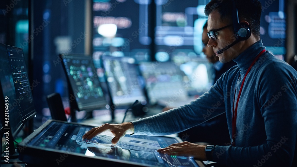 Poster Portrait of Professional IT Technical Support Specialist Working on Computer in Monitoring Control Room with Digital Screens. Employee Wears Headphones with Mic and Talking on a Call.
