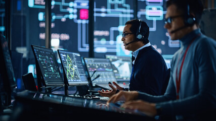 Portrait of Professional IT Technical Support Specialist Working on Computer in Monitoring Control Room with Digital Screens. Employee Wears Headphones with Mic and Talking on a Call.