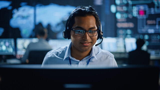 Portrait of Professional IT Technical Support Specialist Working on Computer in Monitoring Control Room with Digital Screens. Employee Wears Headphones with Mic and Talking on a Call.
