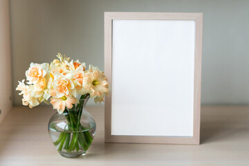 Portrait white picture frame mockup on wooden table. Modern glass vase with Daffodils. White wall background. Scandinavian interior. 
