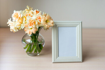 Portrait white picture frame mockup on wooden table. Modern glass vase with Daffodils. White wall background. Scandinavian interior. 