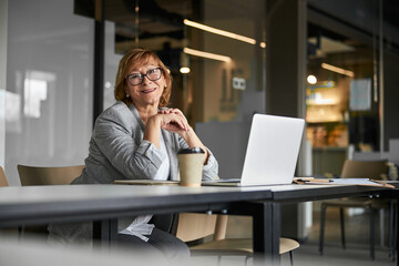 Friendly business lady smiling to camera, sitting near laptop