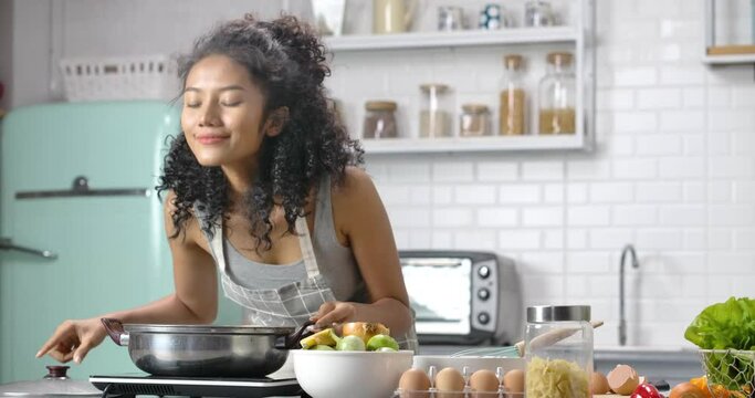 A young, happy and beautiful Asian woman with curly long hair wearing grey gingham apron. She's making a soup for breakfast and open a pot for smelling, scooping up with a smile in the modern kitchen.