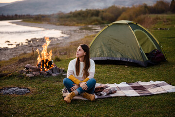 Front view of young brunette in the afternoon sits near a tent and enjoys mountain views. The campfire is lit on the side. Camping and hiking in the mountains.