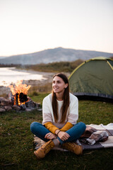 Naklejka na ściany i meble Close up front view of young smiling brunette in the afternoon near a green tent. River, mountains and campfire on background. Camping and hiking in the mountains.