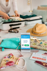 Mobile phone with covid passport with unrecognizable woman preparing suitcase for vacation in the background