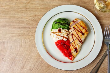 Heart shaped Italian Caprese Salad arranged by Italian basil,buffalo mozzarella and tomatoes look like Italian flag and grilled chicken breast on top with balsamic vinegar on plate with wooden table