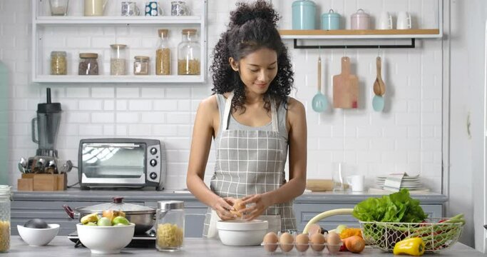 A young, happy and beautiful Asian woman with curly long hair wearing grey gingham apron. She's breaking eggs in a bowl and stir them by whisk, cooking an omelet for breakfast in the modern kitchen.