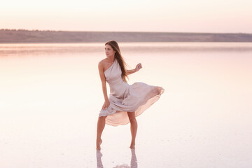 Young blonde woman in an evening airy pastel pink, powdery dress stands barefoot on white...