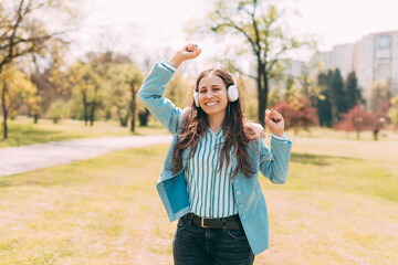 A young smiling woman is dancing happily with her headphones on in park