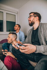 Three happy men playing games and drinking beer while sitting on sofa.