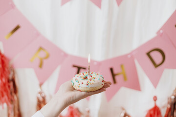 Happy Birthday. Hand holding delicious birthday donut with candle in room with pink garland