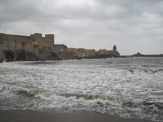 Scenic view of historical castle on the town shore in Collioure, France. Dynamic gray coastline background with storm of the Mediterranean Sea on a cloudy rainy day.
