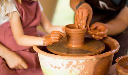 Master class for the child. Teacher hands show to kid how make ceramics dishes on potter wheel. Artist works with clay. Little girl sculpt cup clay